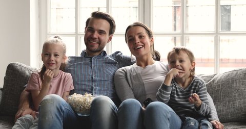 Happy family of four young adult parents mum dad and cute little children watching tv together hold remote control eat snack enjoy television together lounge on sofa having fun in living room at home