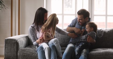 Playful happy young adult parents and funny active small children playing laughing sitting on sofa, cheerful young family mum dad having fun with little kids siblings tickling in living room at home