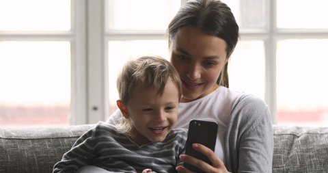 Happy young adult mom teaching little cute kid child son learning using smartphone looking at phone enjoying playing mobile games using apps for tech education entertainment sit on sofa at home