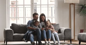 Happy family of four enjoy using smartphone technology relax sit on couch at home, relaxed parents father mother and cute little kids children watch funny social media video on mobile phone together