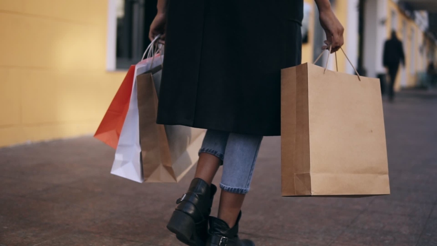  lose up of woman legs wearing stylish black coat and jeans at day time hold colored shopping bags after big shopping day. Fashion shopper shopaholic in slow motion. Rare view