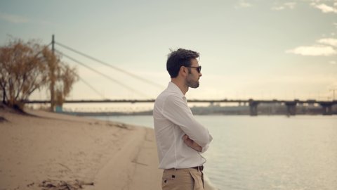 Man Stands On Beach And Looking At Distance. Attractive Businessman In White Shirt Standing Near Bridge And Thinking. Man In Sunglasses Staying And Watching To Horizon. Handsome Man Thinking Something