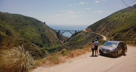 Drobe flying over happy couple near car towards epic Bixby Canyon Bridge and Highway 1 summer panorama in Big Sur USA.