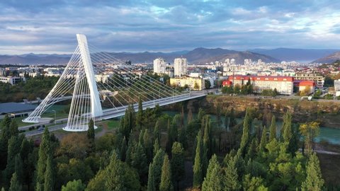 Podgorica, Montenegro: aerial view of the city, featuring Millennium bridge and Moraca river in the morning, at sunrise, under beautiful sky. Cable stayed bridge with green area in the foreground.