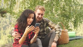 Young couple of tourists watching nasty video on smartphone sitting on picnic in the woods.