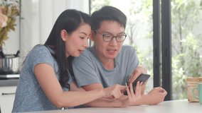 Young Asian couple enjoy shopping online on mobile phone at home. Lifestyle young husband and wife happy buy ecommerce after have breakfast in modern kitchen at house in the morning concept.
