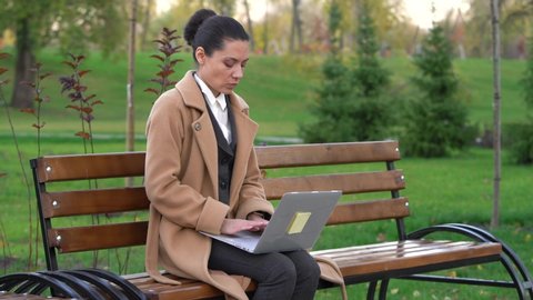 young professional, successful business woman gets good news, cheering celebrating looking at laptop sitting on a wooden park bench among nature on a background of greenery
