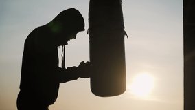 Silhouette of a boxer on a sunset background. It fulfills blows on a pear at high speed. Sweat is flowing from my face. Fatigue. Can't go on. Very hard. Motivational video.