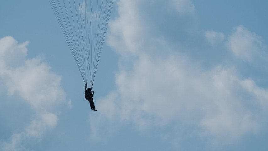 Paratrooper descends slowly on an old torn parachute to ground and flies through sky. Clouds are white. Paratrooper is very brave and desperate down to the ground.