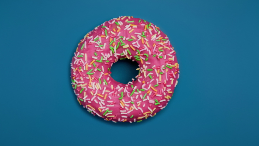 Eating delicious sweet donut with colorful sprinkles on blue background. Timelapse. Bakery and food concept. Top view of colorful frosted doughnut in 4K, UHD | Shutterstock HD Video #1041264898