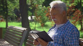 Side view an Elderly man with mustache is reading funny moment in book. Old man is sitting on bench in park and reading ebook - Powered by Shutterstock - Get 15% off with code: PIKWIZARD15