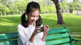 Happy asian teenage girl holding cellphone having fun,female people playing online games on the smartphone while sitting on bench in park,smiling beautiful child girl with enthusiasm plays in phone