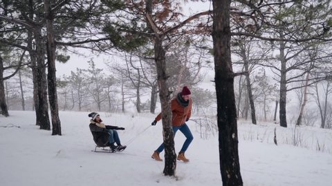 Lovely couple sledding on snowy winter day. Man pull sled with girlfriend on snowfall. Woman have fun and sledge outdoors with boyfriend. People sleigh ride and enjoy Christmas vacation. Slow motion.