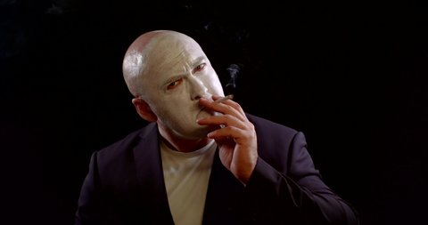 sorrowful and angry actor with white makeup on face is smoking, sitting in black background