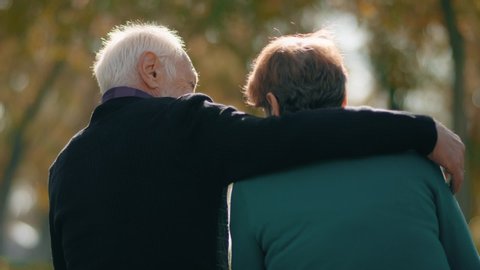 Back view of happy lovely caucasian senior couple sitting in bench and hugging each other . Rear view of romantic old couple sit on bench and enjoy in the open air . Happiness people . Slow Motion .