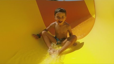 Funny little boy rides off an orange slide in a water park changing position. The boy rides from the slide on his stomach