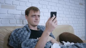 Sick young caucasian man is talking online with doctor getting video consultation showing pills on smartphone sitting on couch under blanket. People and gadgets concept.