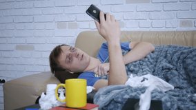 Sick young caucasian man is talking online with doctor getting video consultation showing pills on smartphone lying on couch under blanket. People and gadgets concept.