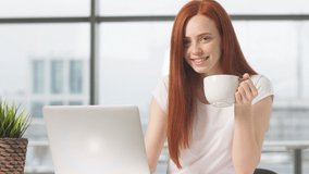 Joyful redhead girl sitting at table with laptop and looking at camera, holding cup of coffee, good start of day