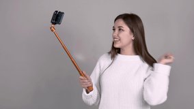 caucasian Girl author of video blog brown haired white jumper long hair on light gray background holds selfiestick with smartphone and emotionally communicates with her subscribers leads live broadcast