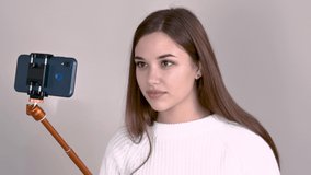 caucasian girl blogger brown haired white jumper long hair on a light gray background holds a selfie stick with a smartphone and communicates with her audience leads a video shoot Close up