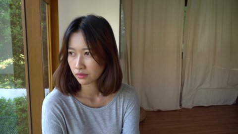4K Depressed young beautiful asian woman face looking at camera. Loneliness woman with sadness eye sitting alone in the room near the window at home. Portrait woman face and mental health care concept
