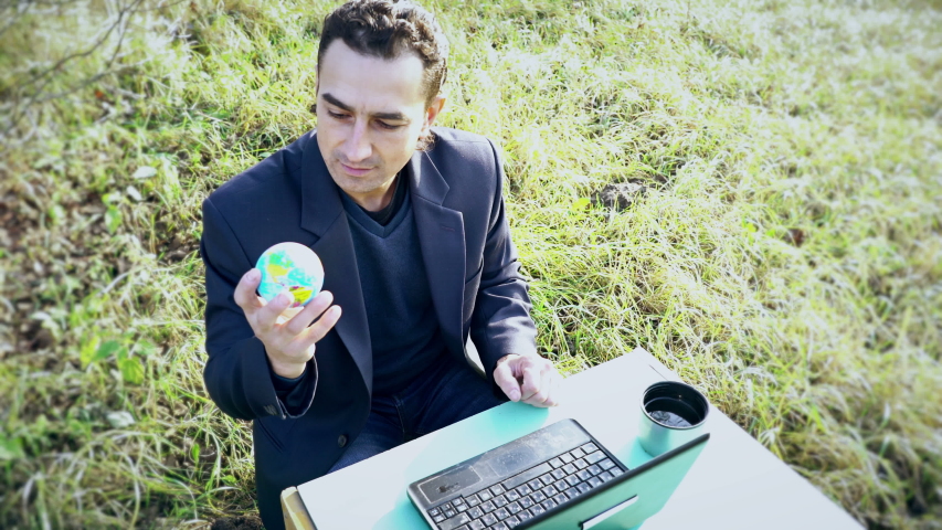Young Attractive Businessman in suit with laptop computer sitting on green meadow outside, office outdoors, big boss holding globe earth and decide human destiny, god hands