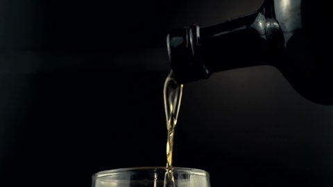 Cognac Pouring In Glass Slow Motion. Golden Whiskey In Goblet Floats On Black Background.Bourbon Pouring In Slow Motion.Running Brandy In Glass From Bottle. Rum Flowing From Brown Bottle.Drink Concept