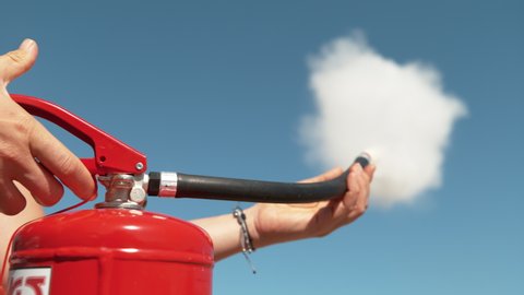 SLOW MOTION, CLOSE UP, DOF: Unrecognizable person squeeze the handle of a fire extinguisher, causing white smoke and foam to come out of the nozzle. Young woman uses a fire extinguisher outdoors.