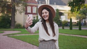 Attractive portrait of pretty slender modern lady in trendy clothes and hat which having video call from her friend while walking in the green lawn in front of cozy dwellings