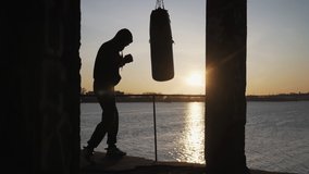 Silhouette of a boxer against the backdrop of a beautiful sunset and a river in an abandoned building, trains blows on a punching bag. Epic video. Motivation. Sports and martial arts.