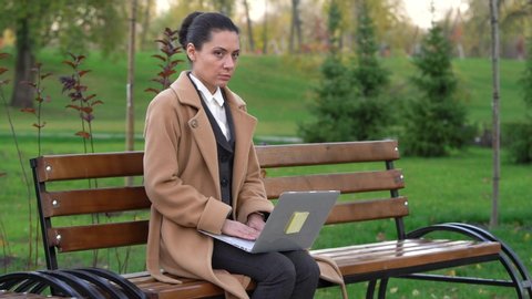 a girl in a coat sits on a wooden bench in the park with a laptop, learns the good news and enjoys it