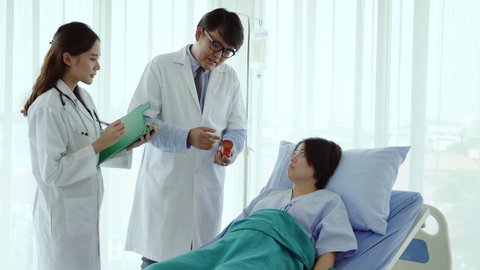 Two Asian doctors standing and talking to patients, a woman lying on the hospital bed Explain medical treatment, health care The use of appropriate drugs for cancer and HIV. Health insurance concept