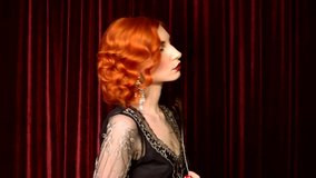 Retro woman with pale skin and red hair in black vintage dress dance against red background in theater. Vintage redhead woman with red lips and retro hairstyle. Noir woman dance with feather