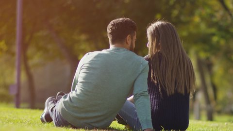 Back view of happy lovely caucasian romantic couple sitting on lawn and hugging each other in park . Rear view of young couple sit on grass and enjoy in the open air. Happiness people . Slow Motion .