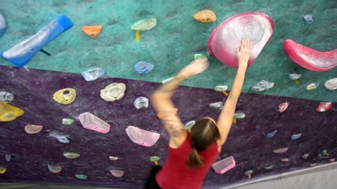 High-angle shot of brunette woman climbing on artificial indoor wall, reaching out, grabbing hand hold with white chalk on it, slipping and landing on floor