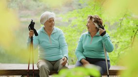Two elderly ladies laugh, talking, relax after a nordic walk in the woods while sitting on a wooden bench. Slow-motion 4k video