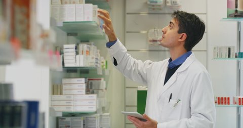 An young male pharmacist consultant is checking with a tablet medicines on the shelf of drug store. 