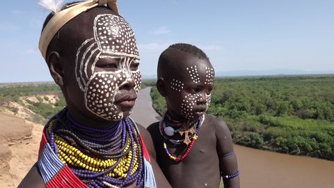 OMO VALLEY, ETHIOPIA – MARCH 2019: Young tribal mother and baby with painted faces and colorful necklaces pose for camera near Omo river in South Ethiopia