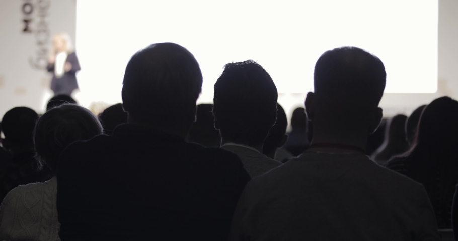 Viewers are listening woman speaker and lookin presentation on business conference in dark, back view. Unrecognizable woman talking microphone in blur background. Educationall training, seminar.