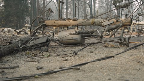 Fallen power pole with transformers attached in Paradise, CA after the Camp Fire in 2018. Power lines fallen across road. Burned trees and homes in background. Dolly out slider shot.