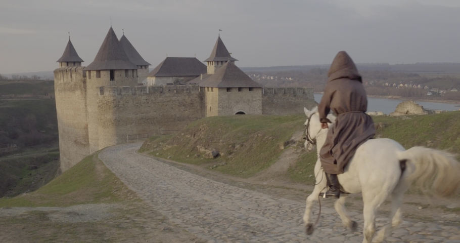 Aerial drone flight view of rider in gray hood cloak on white horse ride into medieval castle gates across bar bridge road. Ancient middle ages stone fortress old atmosphere. Traveler go citadel tower | Shutterstock HD Video #1041431002