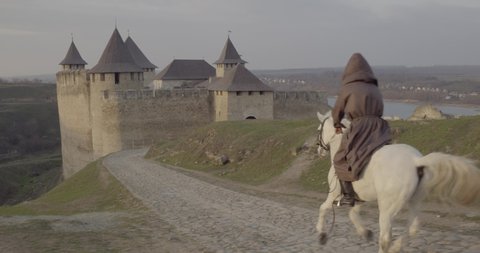 Aerial drone flight view of rider in gray hood cloak on white horse ride into medieval castle gates across bar bridge road. Ancient middle ages stone fortress old atmosphere. Traveler go citadel tower
