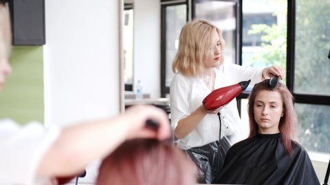 The client's hair is pink. The hair dryer is red. The hairdresser is working in the beauty salon.