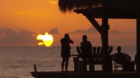 Beautiful sunset on carribean sea. A romantic couple on a wooden jetty looking & shooting the big gold sun over water and orange sky. Dominican Republic, Bayahibe, Playa Dominicus beach