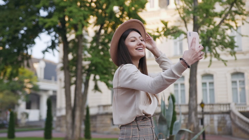 Slow motion of beautiful joyful young woman in stylish clothes and beige hat which strolling in the green yard in front of wonderful houses and photographing on her phone with amusing faces