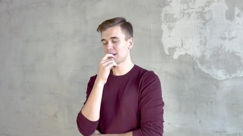Young man in burgundy jumper stands against gray wall bites his lips rubs his chin thinks about solution to problem raises his finger up smiles Medium shot