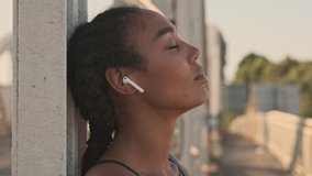 Positive peaceful african american nice woman with earbuds resting during working out on the old bridge