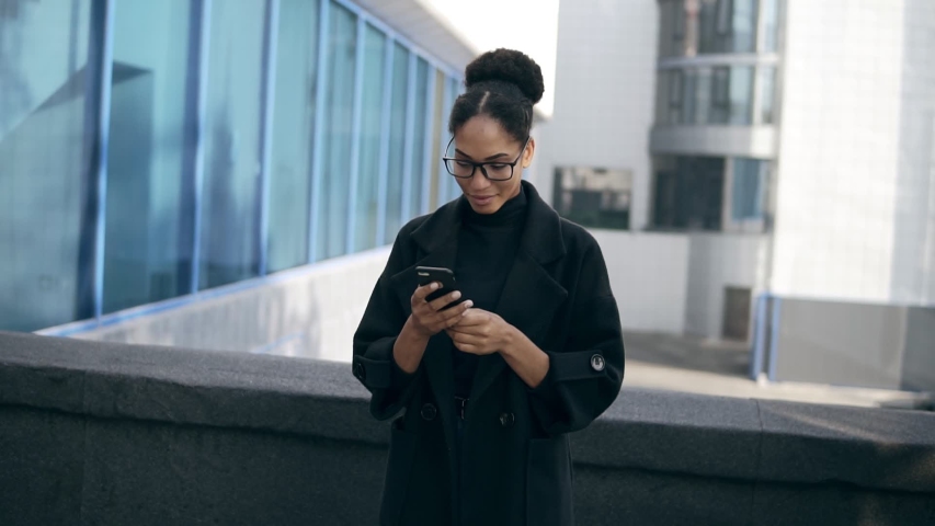 African American woman gots message surprise, reads stunning news on mobile phone. Excited girl starting to gesture, to dance outdoors, on the street. Wearing black coat, jeans and eyeglasses Royalty-Free Stock Footage #1041475918