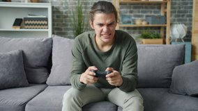 Emotional young man playing video game at home sitting on sofa at home spending leisure time in modern apartment. Youth, lifestyle and fun concept.